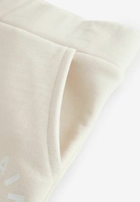 Beige sweatshirts stof med struktureret mønster, der har en foldet ærme og trykte hvide bogstaver på forsiden. Blød og afslappet design.