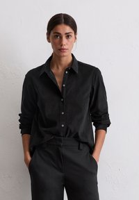 Frau mit dunklem Haar, das nach hinten gebunden ist, trägt ein schwarzes Hemd mit Knöpfen und hochgekrempelten Ärmeln sowie schwarze Hosen vor einer schlichten weißen Wand.