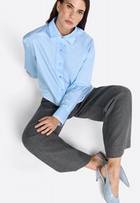 Hellblaues Hemd mit spitzem Kragen, graue weite Hose und blasse blaue Slingback-Absätze, sitzende Haltung mit Hand auf dem Knie.