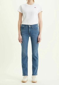 Camiseta de algodón blanca con un pequeño logo rojo, combinada con unos jeans de mezclilla azul claro de corte recto. El modelo lleva zapatillas deportivas color crema.