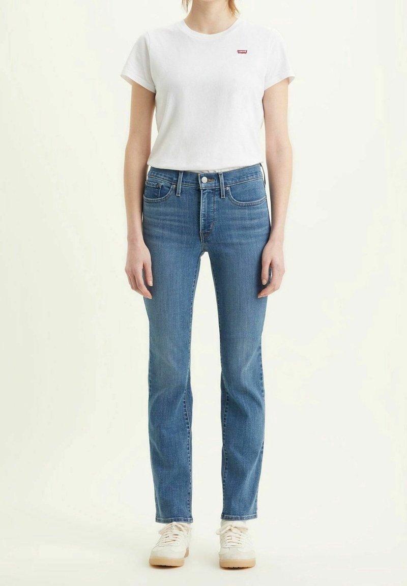 Camiseta de algodón blanca con un pequeño logo rojo, combinada con unos jeans de mezclilla azul claro de corte recto. El modelo lleva zapatillas deportivas color crema.