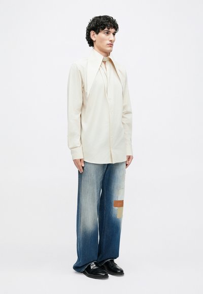 Cream long-sleeve shirt with oversized collar, paired with wide-leg faded denim jeans featuring patchwork accents in tan and beige. Black shoes.