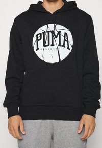 Czarna bluza z kapturem z białą grafiką koszykówki i napisem "PUMA Basketball 1973", noszona z jasnoszarymi spodniami dresowymi.