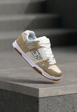 Beige-weißer Sneaker mit Leopardenmuster-Paneelen und weißen Schnürsenkeln schwebt über einem Betonsims vor verschwommenem Treppenhintergrund.