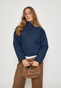 Tumši zils pūkainais mētelis ar augstu apkakli, rāvējslēdzēja aizdari un divām priekšējām kabatām. Modelis tur rakstainu brūnu austu somu.