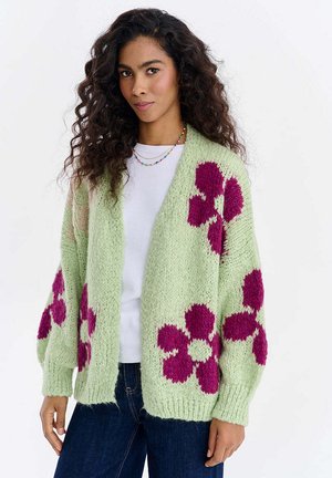 Kobieta z długimi, kręconymi włosami nosi jasnozielony sweter z dzianiny z dużymi fioletowymi wzorami kwiatowymi, białą koszulę i ciemnoniebieskie dżinsy.