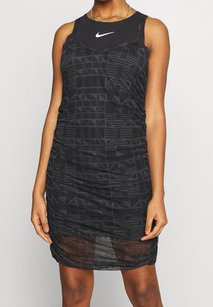 Frau trägt ein schwarzes ärmelloses Kleid mit geometrischen Mustern und ein mehrlagiges Nike Tanktop mit weißem Swoosh-Logo.