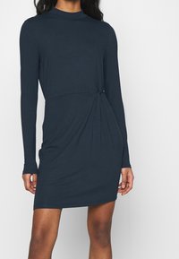 Mörk marinblå långärmad klänning med hög halsringning och vriden knutdetalj i midjan; tillverkad av mjukt, elastiskt tyg med en slät yta.
