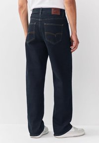 Mand iført mørkeblå lige jeans og hvide sneakers, står mod en ensfarvet lys baggrund.