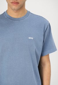 Világoskék, rövid ujjú póló, puha pamutból készült, kerek nyakkivágással és egy kis fehér "obey" logóval a bal mellrészen.