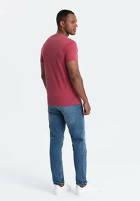 Camiseta roja de manga corta, ligeramente ajustada, combinada con jeans azules. El modelo lleva zapatillas blancas y está posicionado en un ángulo trasero.