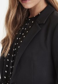 Czarna marynarka o gładkiej fakturze, z klasycznym krojem i wyciętymi klapami, zestawiona z czarną bluzką ozdobioną białymi groszkami.