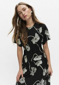 Młoda kobieta z długimi brązowymi włosami, ubrana w czarną sukienkę w białe kwiatowe wzory oraz srebrne kolczyki w kształcie kół, na jednolitym jasnym tle.