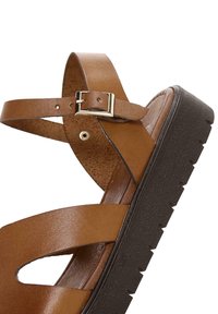 Sandale en cuir marron avec boucle dorée et semelle noire épaisse texturée, dotée de larges brides et d'une bride de cheville ajustable.