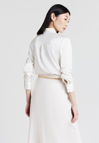 Donna con capelli neri lisci che indossa una blusa di raso bianca e una gonna coordinata con una cintura sottile beige, rivolta di lato contro uno sfondo neutro.