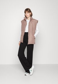 Colete acolchoado sem mangas em castanho claro, com colarinho alto e fecho de zíper, combinado com calças largas pretas e ténis multicoloridos.