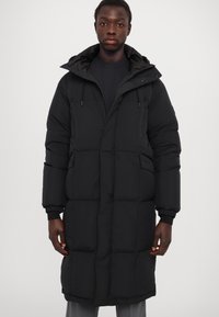 Abrigo tipo puff negro con capucha, con un diseño acolchado, mangas largas y puños elásticos. El material parece grueso y aislante.