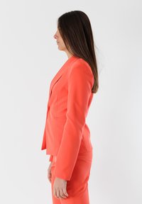 Blazer corallo da donna con maniche lunghe, vestibilità sartoriale, rever a punta e texture liscia. Le maniche a campana e i pantaloni abbinati completano il completo.