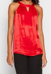 Haut halter en satin rouge avec un décolleté en trou de serrure et un détail froncé au col ; texture lisse et coupe légèrement ample.