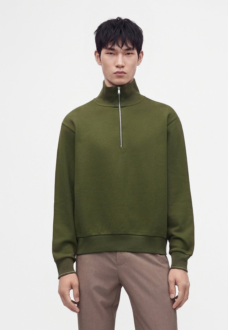 Homme portant un pull à manches longues vert olive avec col à fermeture éclair partielle et pantalon marron clair, debout devant un fond blanc uni.