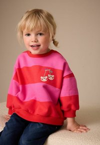 Sudadera a rayas rojas y rosas con diseño bordado de cerezas. Tejido suave, ajuste holgado, puños y cuello acanalados. Combinada con vaqueros de mezclilla.