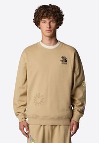 Beige sweatshirt med långa ärmar, ribbad mudd och nederkant. Har broderad logotyp och färgglada grafiska mönster på ärmarna och framsidan.