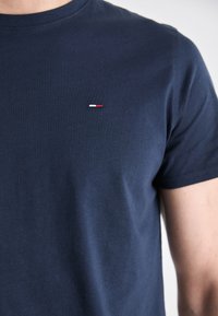 Uomo che indossa una maglietta blu navy semplice con un piccolo logo rettangolare rosso, bianco e blu sul lato sinistro del petto.
