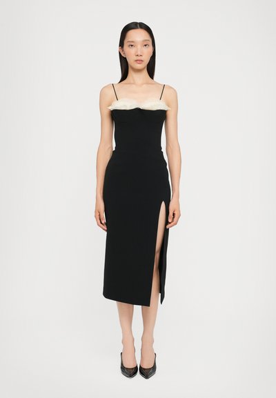 Black fitted dress with a white feathered accent at the bust, featuring thin straps and a thigh-high side slit. Worn with black pointed heels.