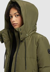 Young person wearing an olive green hooded puffer jacket with black zipper and snap buttons, facing slightly left on a gray background.