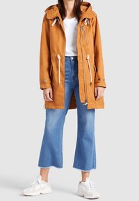 Pomarańczowa lekka parka z kapturem, regulowanymi sznurkami, zamkiem z przodu i kieszeniami. Do tego wyblakłe niebieskie dzwony i białe trampki.