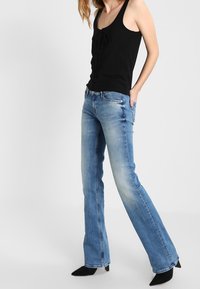 Nainen, joka pukeutunut mustaan hihattomaan toppiin ja vaaleansiniset bootcut-farkut, jalassaan mustat korkokengät, seisoo käsi taskussa.