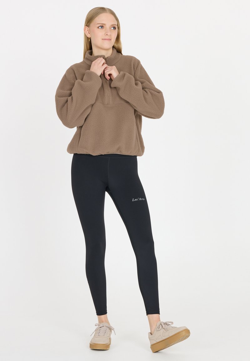 Brauner Fleece-Pullover mit hohem Kragen, kombiniert mit schwarzen Leggings mit weißem Text. Helle graue Sneaker mit hellbrauner Sohle runden den Look ab.