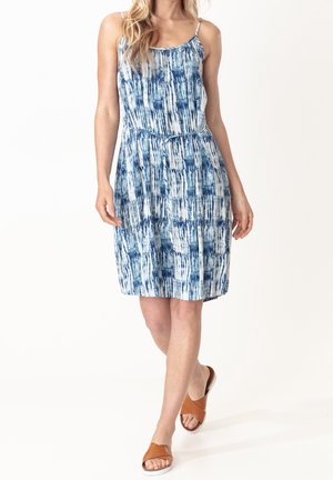 Frau trägt ein ärmelloses, knielanges Kleid mit blau-weißem Muster und dünnen Trägern, dazu braune flache Sandalen mit offenen Zehen, geht nach vorne.