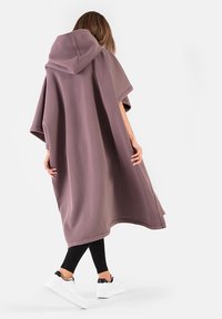 Kapturowy poncho z miękkiej tkaniny w kolorze delikatnego mauve, luźny krój z krótkimi rękawami. Posiada gładki dół i swobodny fason, zestawione z czarnymi legginsami i białymi trampkami.
