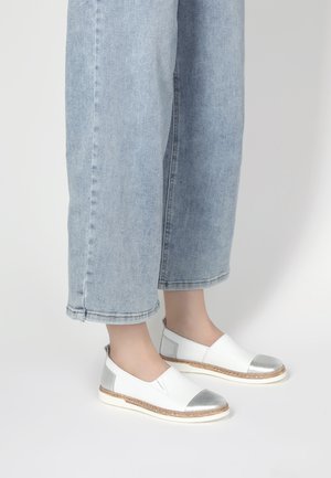 Weiße Slipper mit einer silbernen Zehenkappe und Akzenten, strukturiertem geflochtenem Jutemittelsohle und einem glatten Lederoberteil, vor hellblauen Jeans.