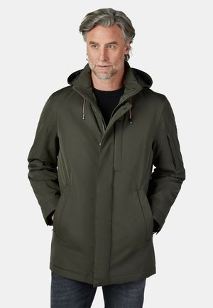 Mann mit grauen Haaren, der eine dunkelgrüne Kapuzen-Winterjacke trägt und die Hände in den Taschen hat, schaut leicht zur Seite vor weißem Hintergrund.