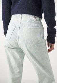 Svetlo modré džíny s vyblednutou textúrou, vysokým pásom a zadnými vreckami. Na pásku je značka Calvin Klein Jeans.