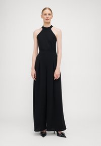 Black halter jumpsuit with a fitted bodice and wide-legged pants. Features pleats at the waist and is complemented by pointed black heels.