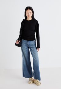 Black long-sleeve top with gathered design, paired with wide-leg blue denim jeans. Model wears beige clogs adorned with decorative studs.