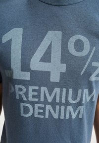 Blå bomull T-skjorte med stor hvit tekst som leser "14% PREMIUM DENIM." Teksten er fet og sentrert, med en myk, glatt tekstur.