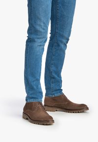 Botas de ante marrón hasta el tobillo con punta redonda, diseño de cordones y parte superior texturizada, combinadas con jeans ajustados azules. Suelas de goma antideslizantes.