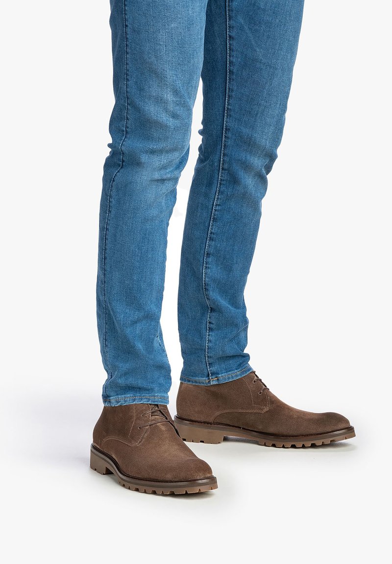 Botas de ante marrón hasta el tobillo con punta redonda, diseño de cordones y parte superior texturizada, combinadas con jeans ajustados azules. Suelas de goma antideslizantes.