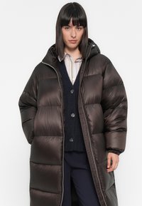Vrouw in een lange, zwarte pufferjack over een marineblauwe cardigan en gestreept shirt, staande tegen een effen witte achtergrond.