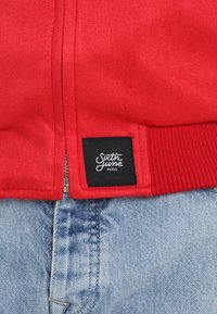 Rode jas met een gladde textuur, voorzien van een geribde kraag en manchetten. Het label zegt "Sixth June Paris" op een zwarte achtergrond.