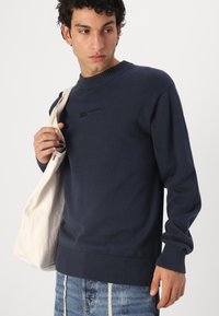 Pulover tricotat de culoare bleu marin, cu guler și manșete rib, având un logo brodat negru. Modelul ține o geantă de umăr de culoare crem.