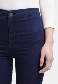 Vysoké tmavě modré džíny s hladkou texturou, s kovovým knoflíkem a šitými švy, přiléhající v oblasti pasu a boků.