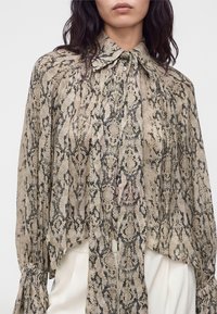 Femme portant un chemisier ample à manches longues avec un imprimé serpent beige et noir et un col noué, associé à un pantalon blanc.