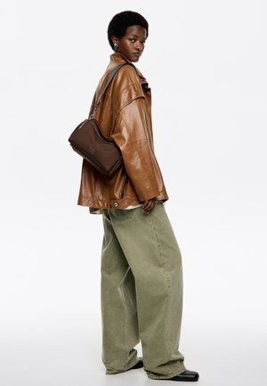 Brązowa skórzana kurtka oversize, zielone szerokie spodnie, a także mała brązowa torebka na ramię z zamkiem. Czarny botki za kostkę dopełniają ten wygląd.