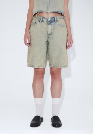 Person trägt hellblaue, knielange Jeansshorts, weiße Socken, schwarze Slip-On-Schuhe und ein kurzes Jeans-Top.