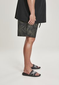 Fekete póló, sötétzöld és fekete mintás short, pálmalevél mintával, és fekete papucs, amelyen fehér "URBAN" felirat található.
